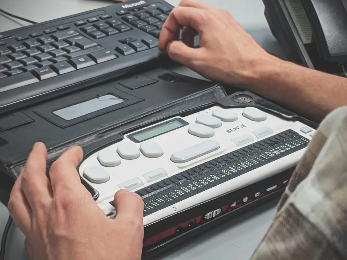 Vak férfi braille képernyőolvasó eszközt használ egy íróasztalnál — ez az a segítő technológia, amelynek támogatására az ARIA készült.